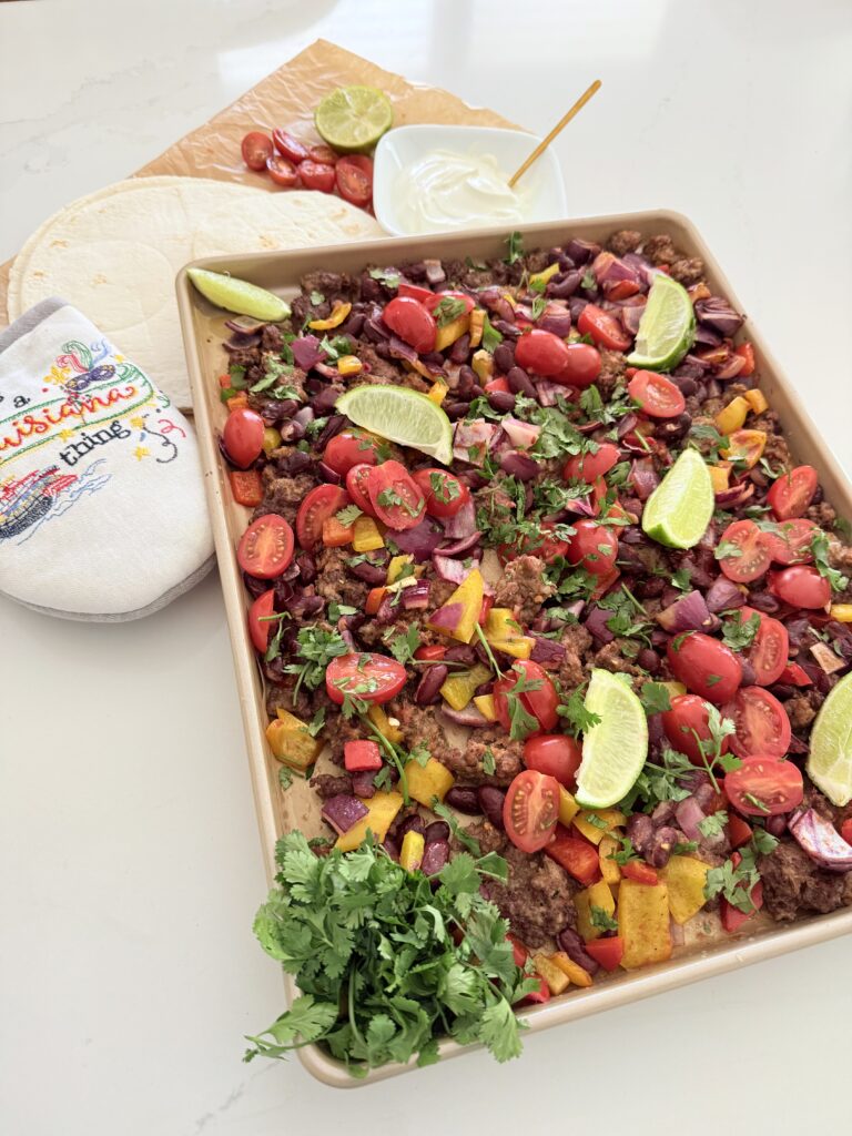 Cooked chopped red onion, garlic cloves, chopped red and yellow bell pepper, kidney beans, ground spicy pork sausage, ground beef, cajun seasoning, ground cumin, dried oregano, tomato paste in a tube, fresh cilantro, limes cut into wedges, and cherry tomatoes on a sheet pan on the counter. An oven mit and tortillas, sour cream in a small white bowl and cut cherry tomatoes and a lime half in the background on the counter.