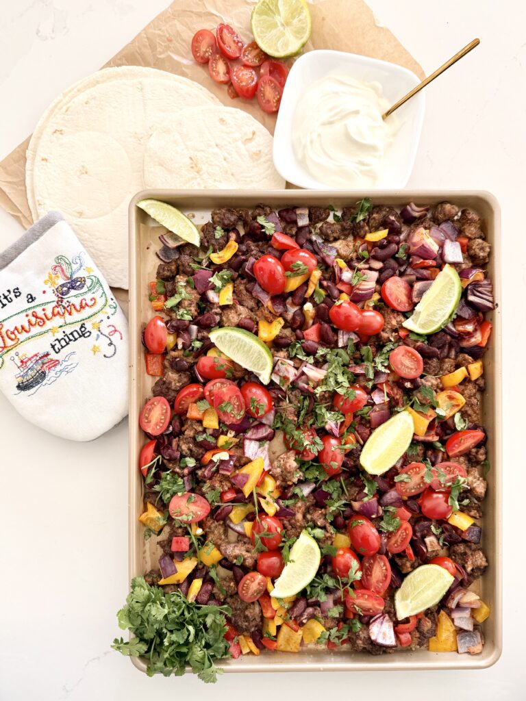 Cooked chopped red onion, garlic cloves, chopped red and yellow bell pepper, kidney beans, ground spicy pork sausage, ground beef, cajun seasoning, ground cumin, dried oregano, tomato paste in a tube, fresh cilantro, limes cut into wedges, and cherry tomatoes on a sheet pan on the counter. An oven mit and tortillas, sour cream in a small white bowl, cut cherry tomatoes, and a lime half in the background on the counter.