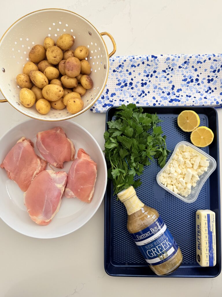 Skinless chicken thighs in a white shallow bowl, baby Dutch yellow potatoes in a white and gold strainer, on a dark blue sheet pan is a handful of parsley, Greek salad dressing, crumbled feta cheese in a plastic container, a stick of butter, I have lemon and above that is a blue and white hand towel on the counter.