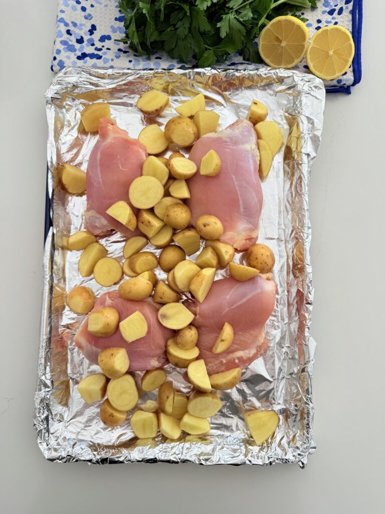 On a line baking sheet is the four chicken thighs and halved cut mini yellow Dutch potatoes on the counter.