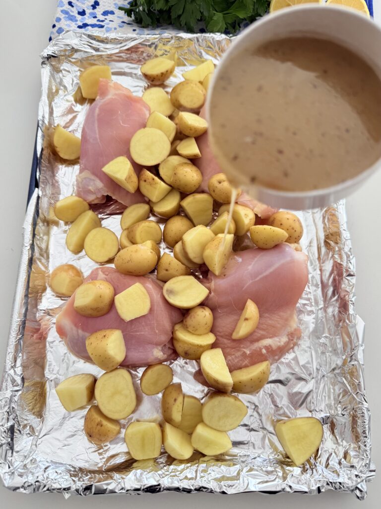 On a line baking sheet is the four chicken thighs and halved-cut mini yellow Dutch potatoes with Greek salad dressing being poured on top on the counter.