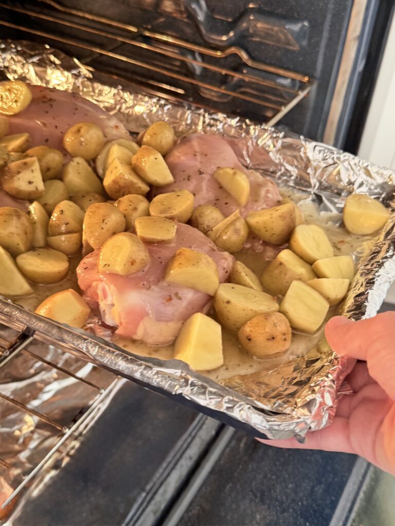 On a line baking sheet are the four chicken thighs and halved-cut mini yellow Dutch potatoes with a tab or half a tablespoon of butter on each of the skinless chicken thighs being put into a oven.