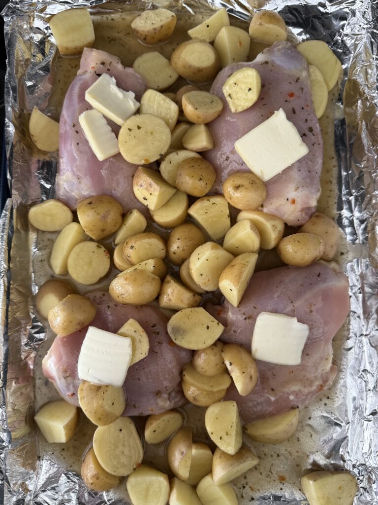 On a line baking sheet are the four chicken thighs and halved-cut mini yellow Dutch potatoes with a tab or half a tablespoon of butter on each of the skinless chicken thighs on the stove top.