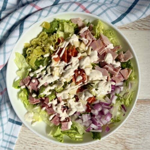 Fresh homemade Italian salad with lettuce, diced ham, salami, pepperoni, chopped onions, and creamy salad dressing on a white bowl with a blue checkered cloth.