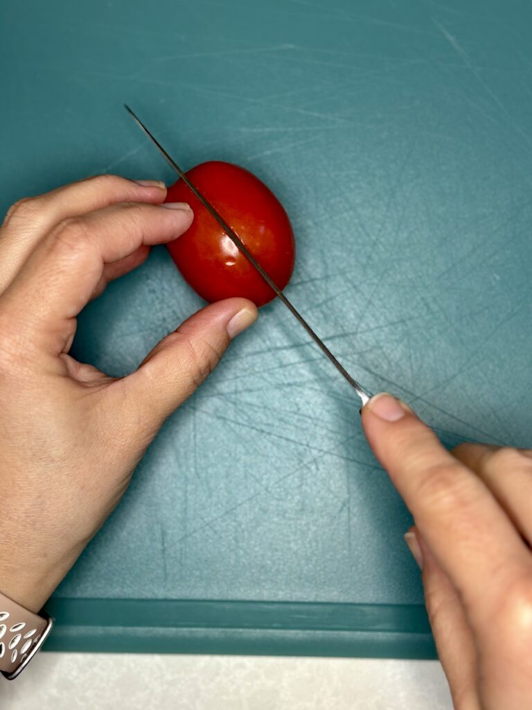 a tomato being cut in 2 on a cutting board with a knife