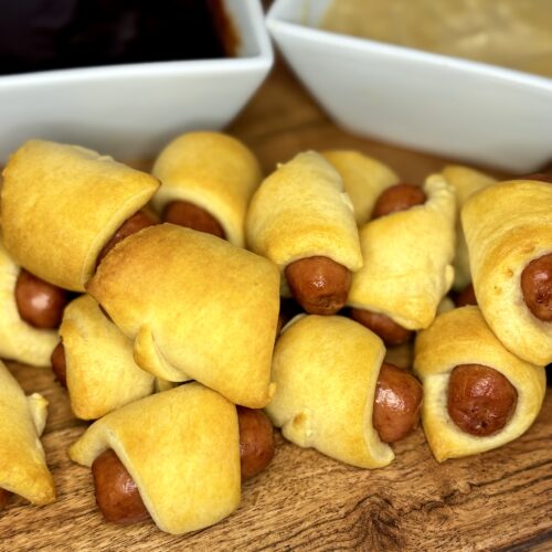 cooked pigs in a blanket on a board with dipping sauces