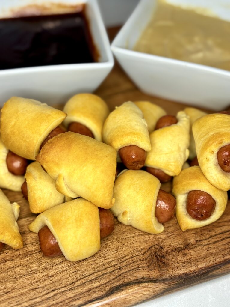 cooked pigs in a blanket on a board with dipping sauces