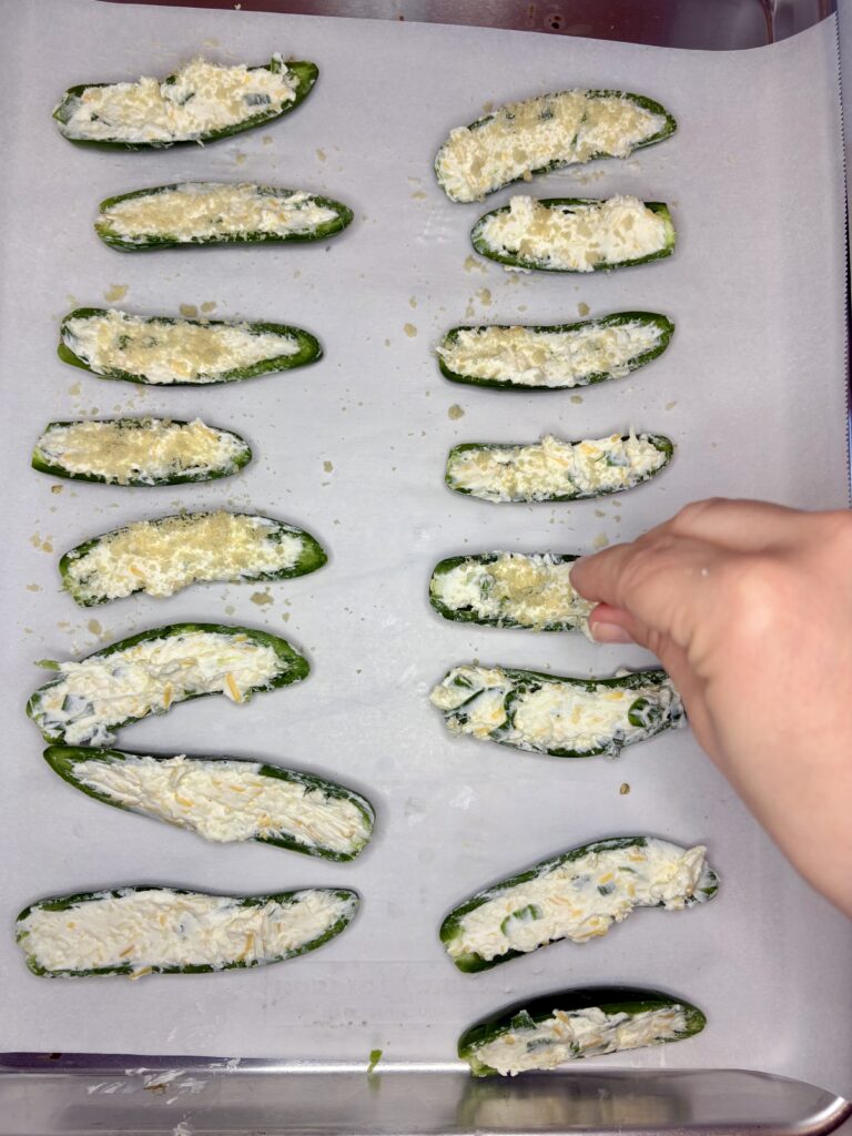 jalapeno peppers filled with cream cheese mixture on a baking sheet lined with parchment paper being sprinkled with panko bread crumbs