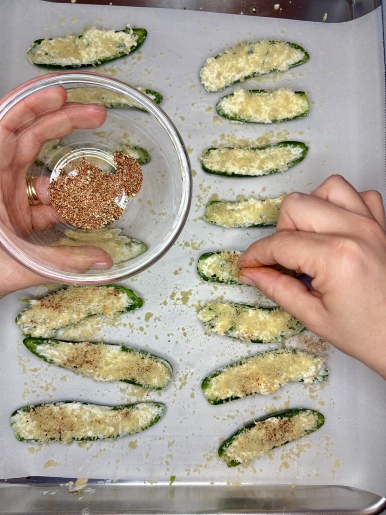 jalapeno peppers filled with cream cheese mixture topped with panko bread crumbs on a baking sheet lined with parchment paper being sprinkled with seasoning