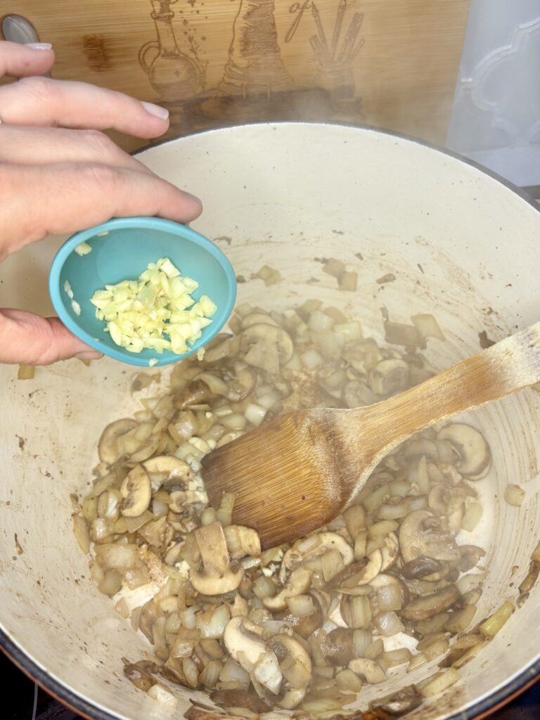 garlic being added to mushrooms and diced onions in a pot on a stove