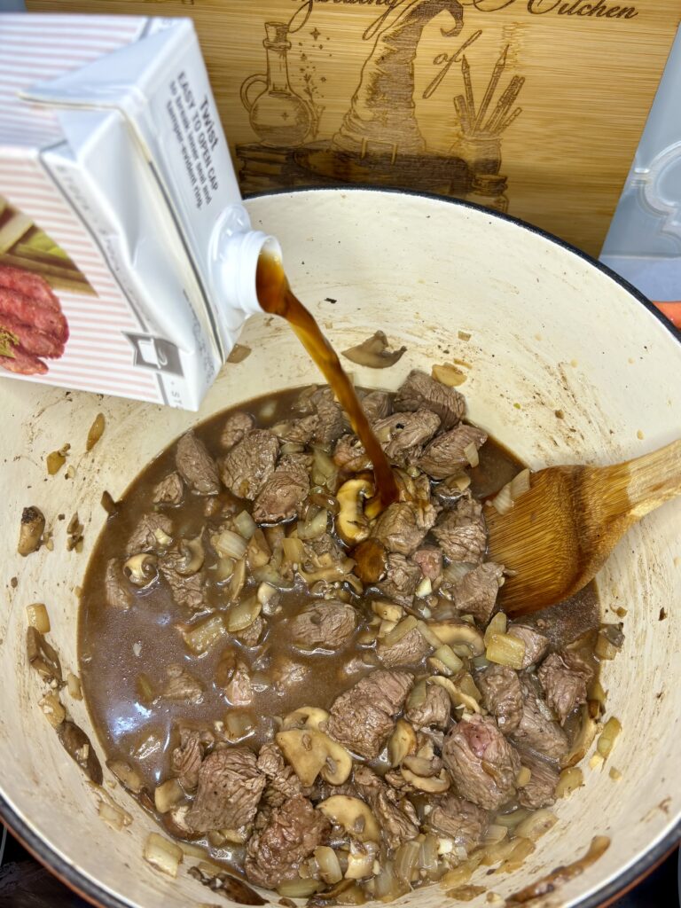 beef stock being poured into pot with beef, onion, mushrooms, garlic and brandy 