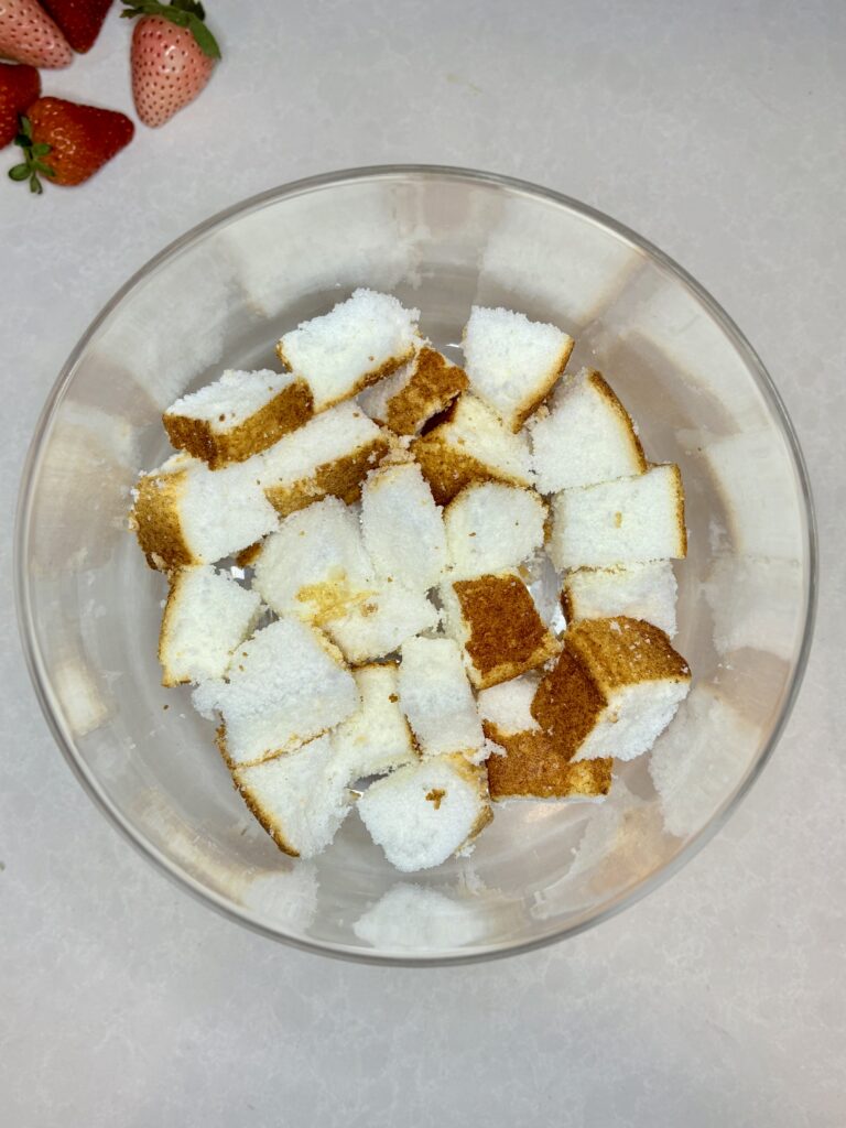 cubed angel food cake in the bottom of a trifle dish on a counter next to strawberries