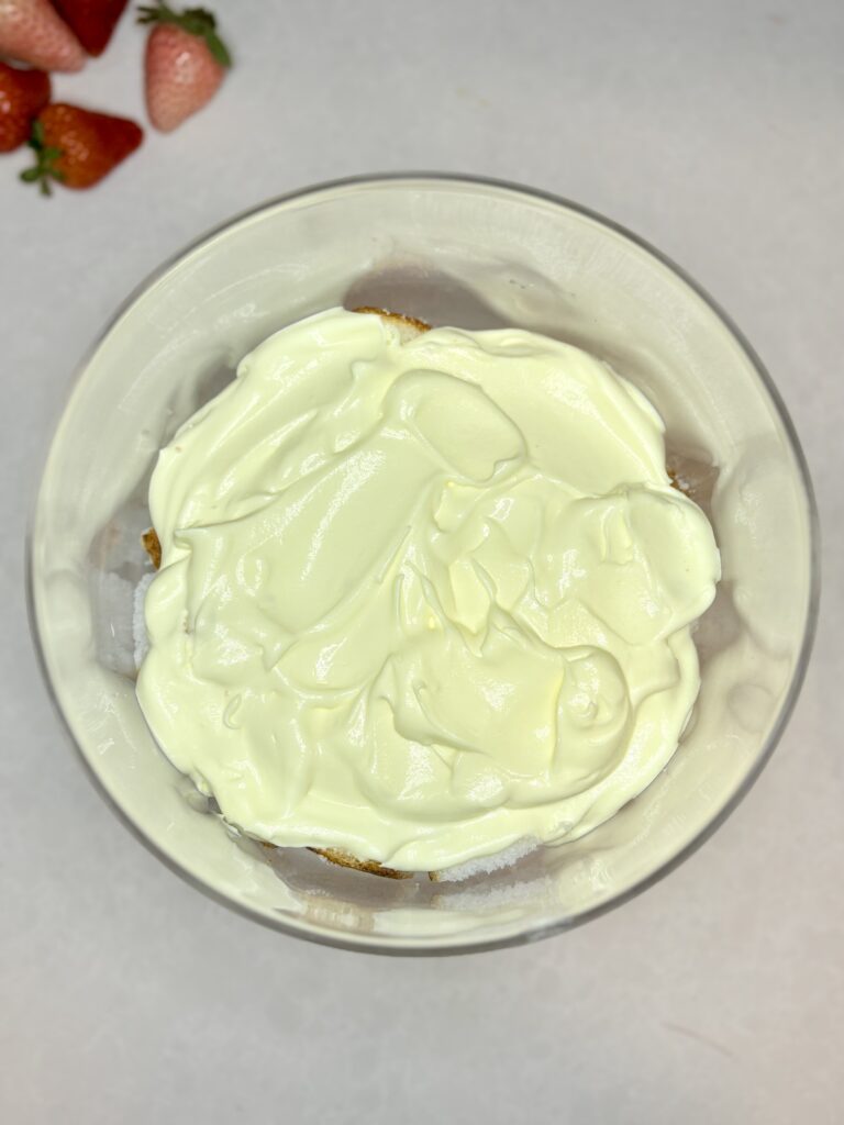 pudding spread over cubed angel food cake in the bottom of a trifle dish on a counter next to strawberries