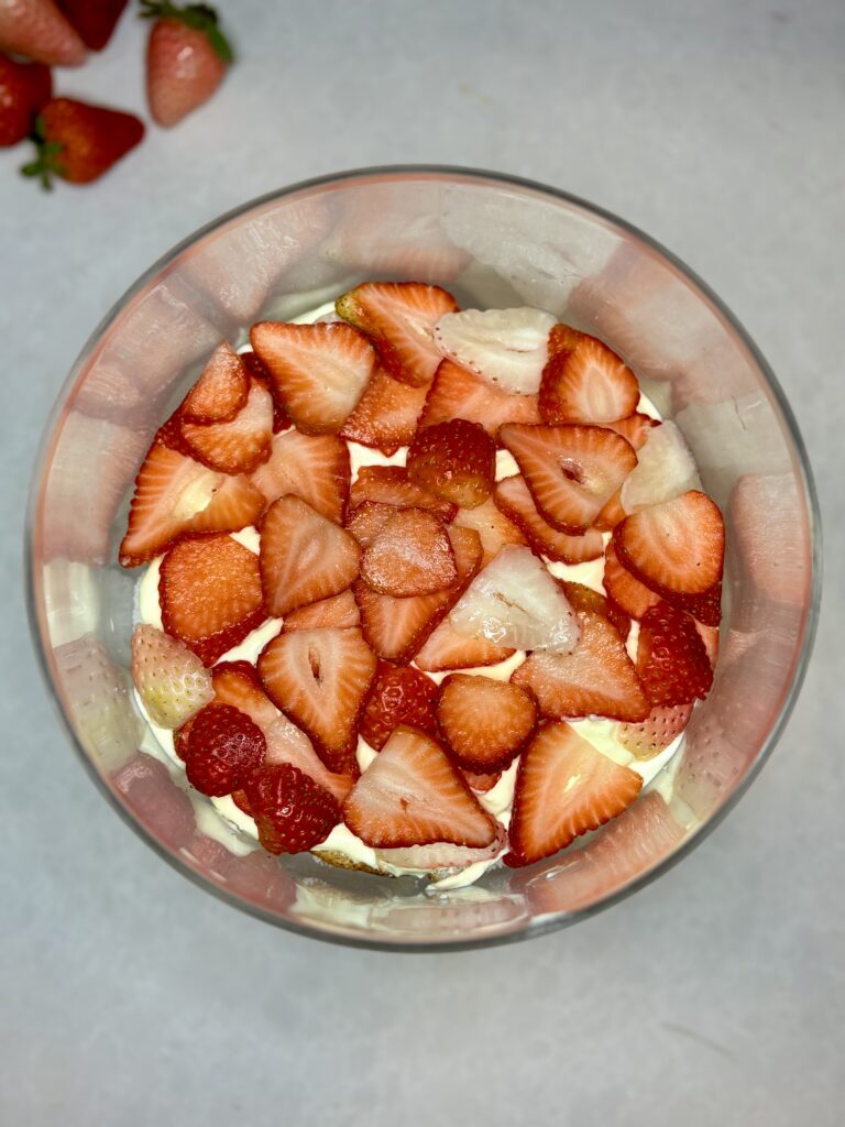 sliced strawberries layered over pudding and angel food cake in a trifle dish on a counter next to strawberries