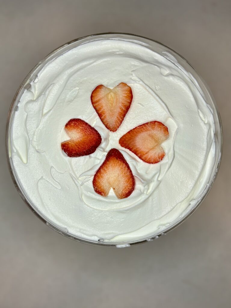 the top of a trifle dish filled with pudding, angel food cake cubes, and strawberries covered in whipped topping with 4 strawberry slices on top