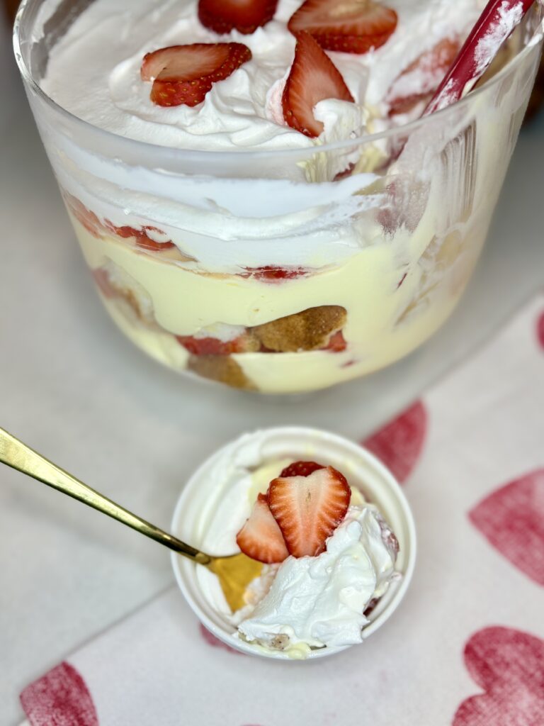 a bowl with a serving of strawberry vanilla trifle and a gold spoon with a heart covered towel next to the trifle dish