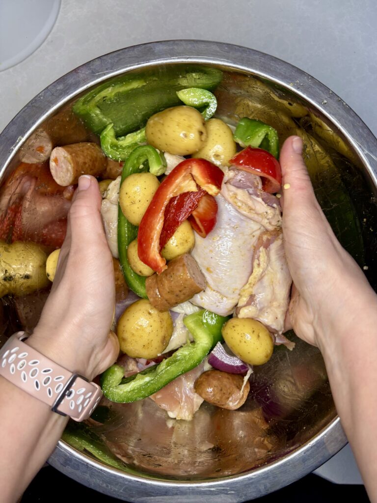 hands tossing chicken, sausage, and vegetables in a bowl with oil and seasoning