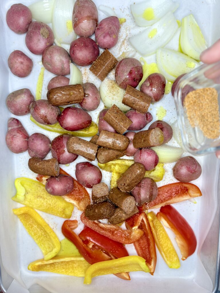 sausage and vegetables on a sheet pan with seasoning being sprinkled on 