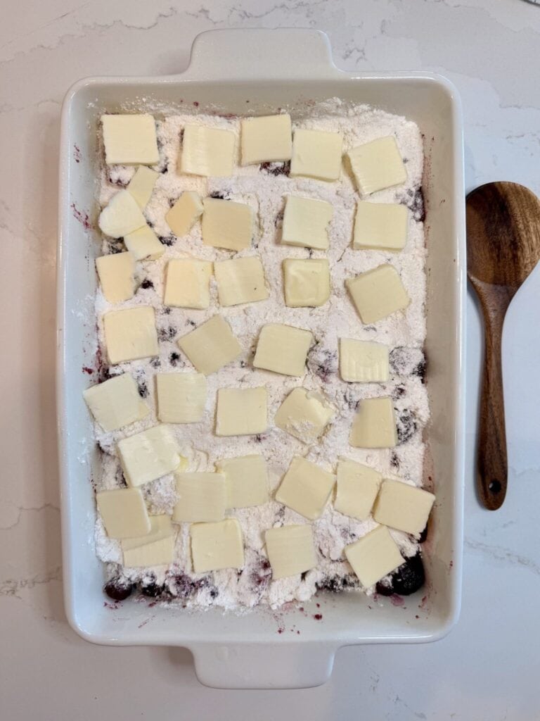 Mixed berries with sugar and ginger mixed together and a white baking dish on the white counter with a brown spoon beside it with the white cake. Mix on top and square pieces of cut butter placed on top.