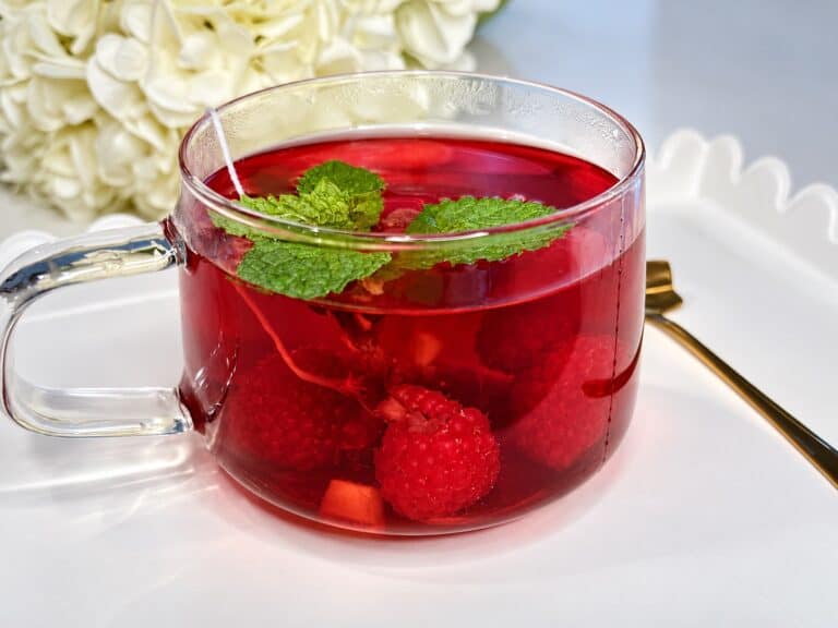 Refreshing raspberry ginger tea in a clear glass cup with fresh mint leaves, served on a white surface with a gold spoon nearby.
