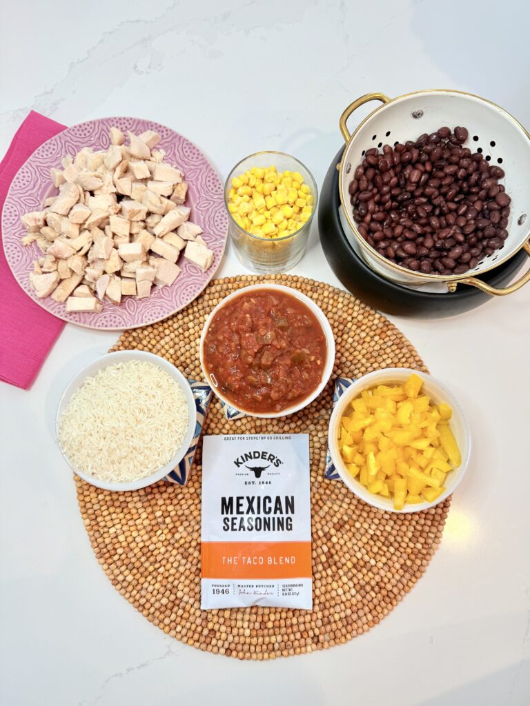 A packet of taco Mexican seasoning blend, white rice in a bowl, jar of salsa in a small bowl, chopped up and diced yellow bell pepper in a small bowl, frozen corn in a glass, chicken that is prepackaged store-bought chicken, cut up into bite side pieces and drained, black beans all on the counter.