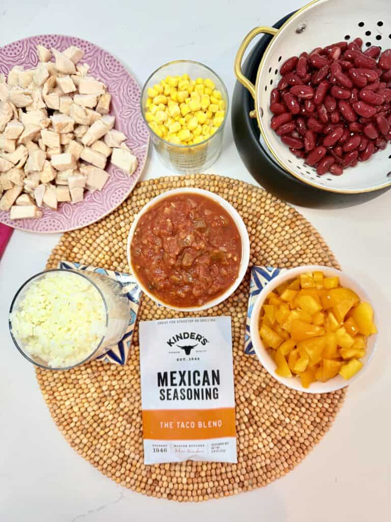 A packet of taco seasoning, jar of salsa and a small bowl, frozen cauliflower rice in a glass jar, drained kidney beans in a strainer, frozen corn in a glass, orange diced bell pepper, and prepared diced chicken on a purple plate. On the counter.