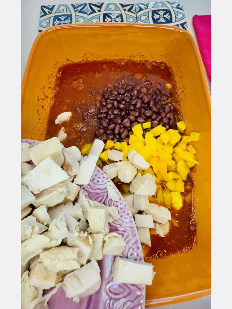 Adding diced pieces of chicken and the chopped yellow bell pepper and the drained black beans to the 2 cups of water, the salsa, and the  taco seasoning blend in an orange casserole dish on the counter.