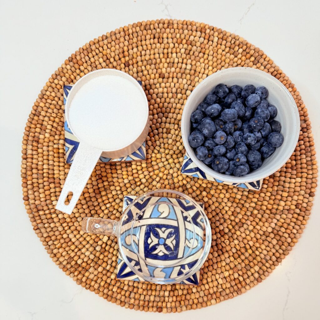 Water in a glass, fresh blueberries in a small bowl, sugar, and a measuring cup on a brown placemat on the counter.