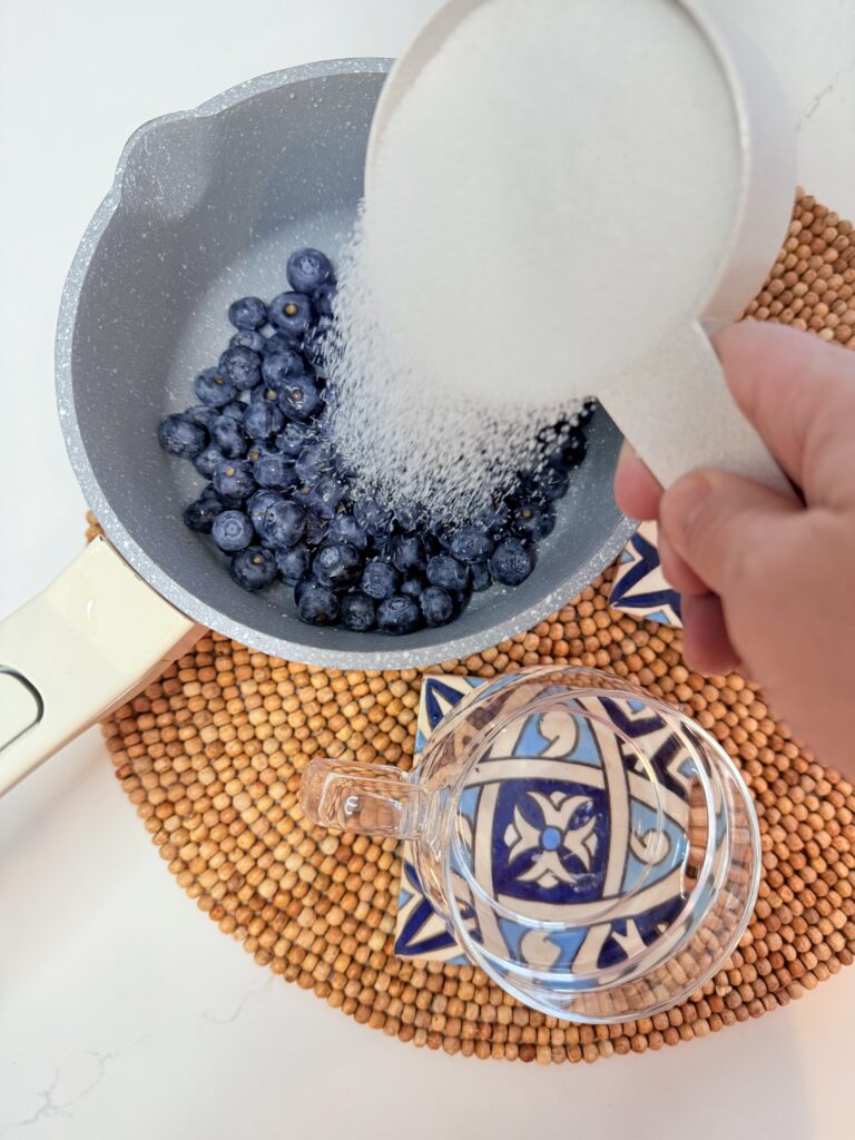 Pouring sugar over fresh blueberries in a pot on a brown mat on the counter.