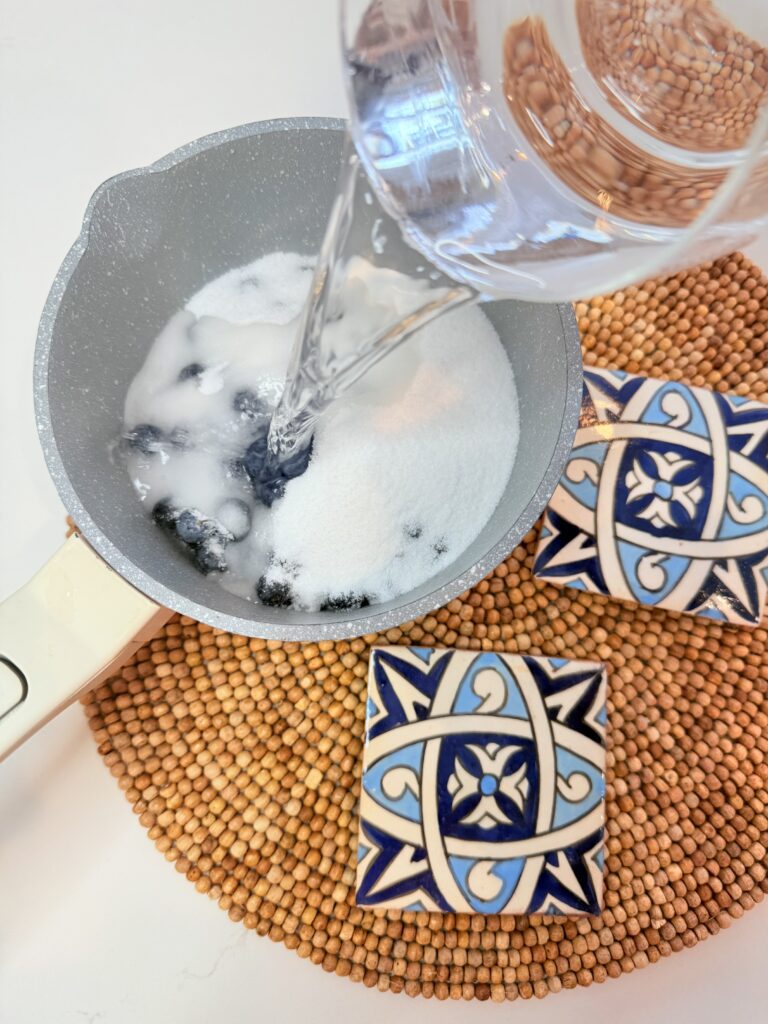 Pouring water over sugar and fresh blueberries in a pot on a brown mat on the counter.