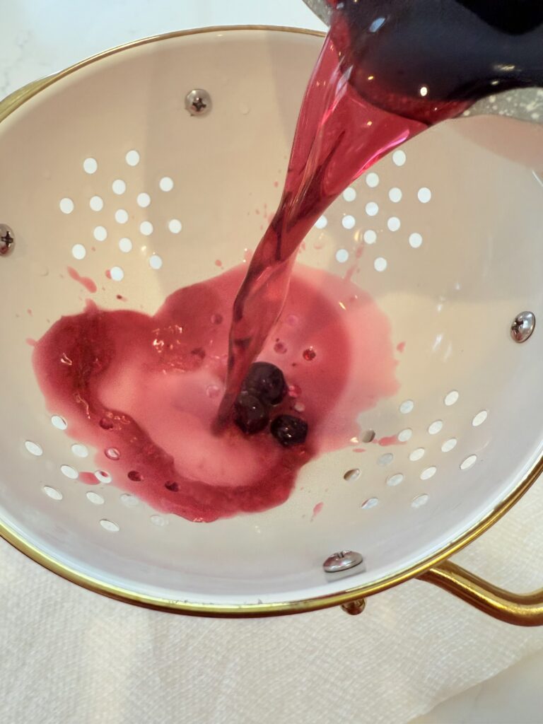 Cooled blueberry simple syrup being strained in a white strainer into a glass container on the counter.