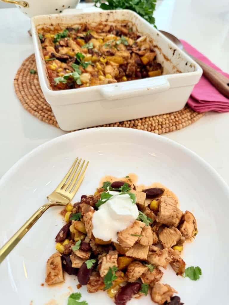 Low-carb taco chicken casserole in a white casserole dish in the background and in the foreground is a piece of the low-carb taco chicken casserole on a white plate with a gold fork with sour cream developed on top with cilantro sprinkled on top all on the counter.