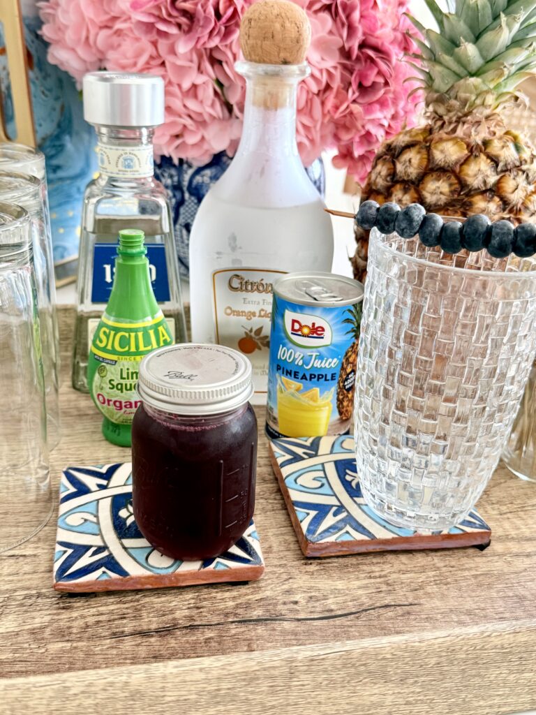 Tequila, blueberry simply syrup, lime juice, pineapple juice in a can, orange liqueur, and fresh pineapple on a brown tray on the counter. As well as blueberries on a glass shaker on the tray on the counter. On a brown tray