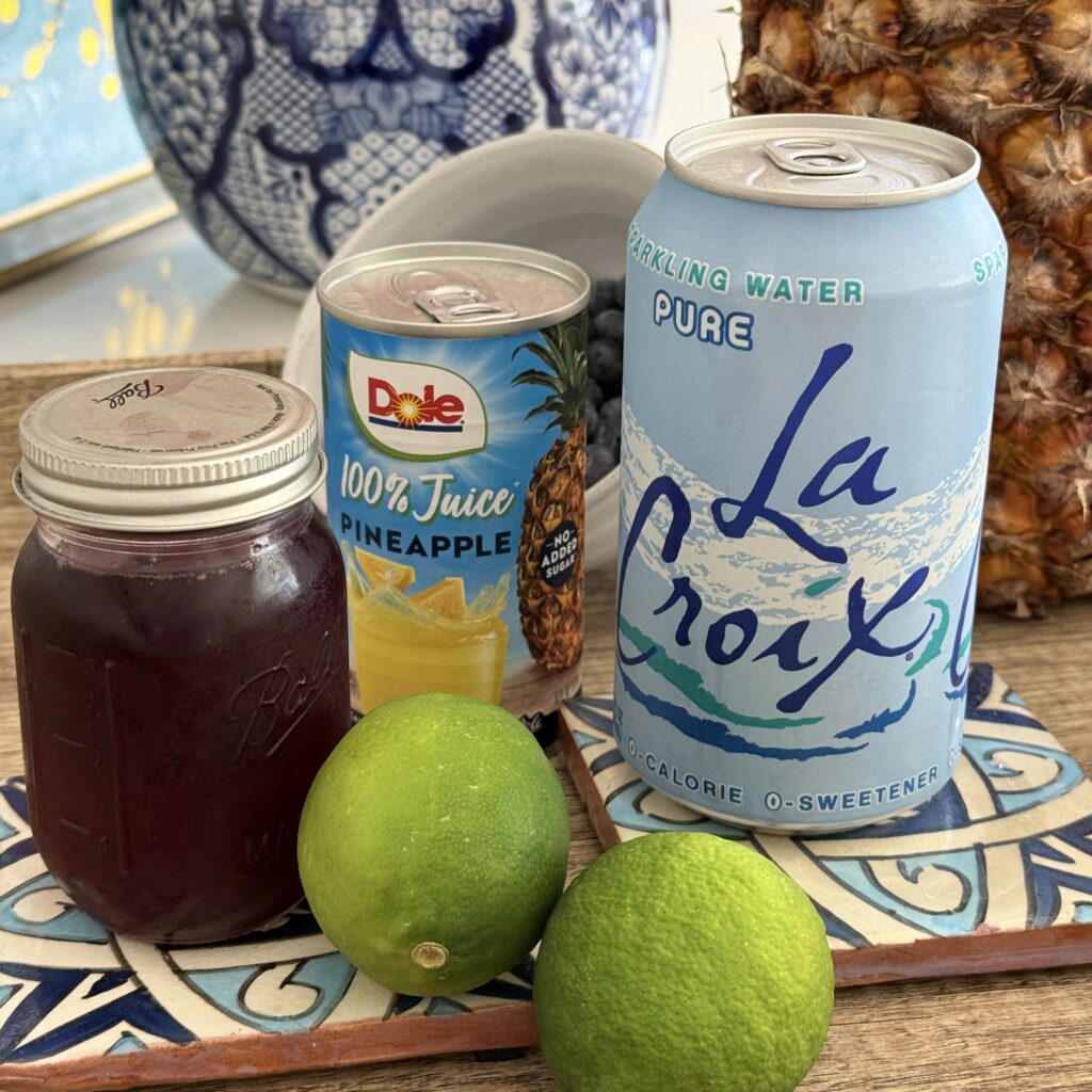 Canned pineapple juice, blueberry simple syrup, two limes, blueberries in a dish in the background, and unflavored sparking water in a blue can and a brown tray on the counter.