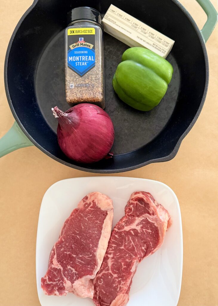 2 Strip Loin Steaks; 10 oz each on a white plate, and Montreal Steak seasoning, butter, green bell pepper and red onion in a black cast-iron skillet all on top of brown parchment paper on the counter.