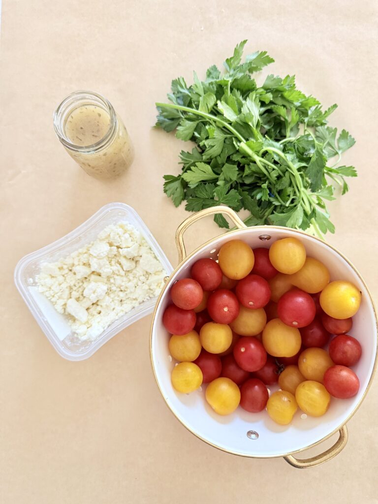  The salad dressing is in a small glass jar, the feta cheese is in a square container, and a handful of parsley and yellow and red cherry tomatoes are in a white strainer on brown parchment paper on the counter.