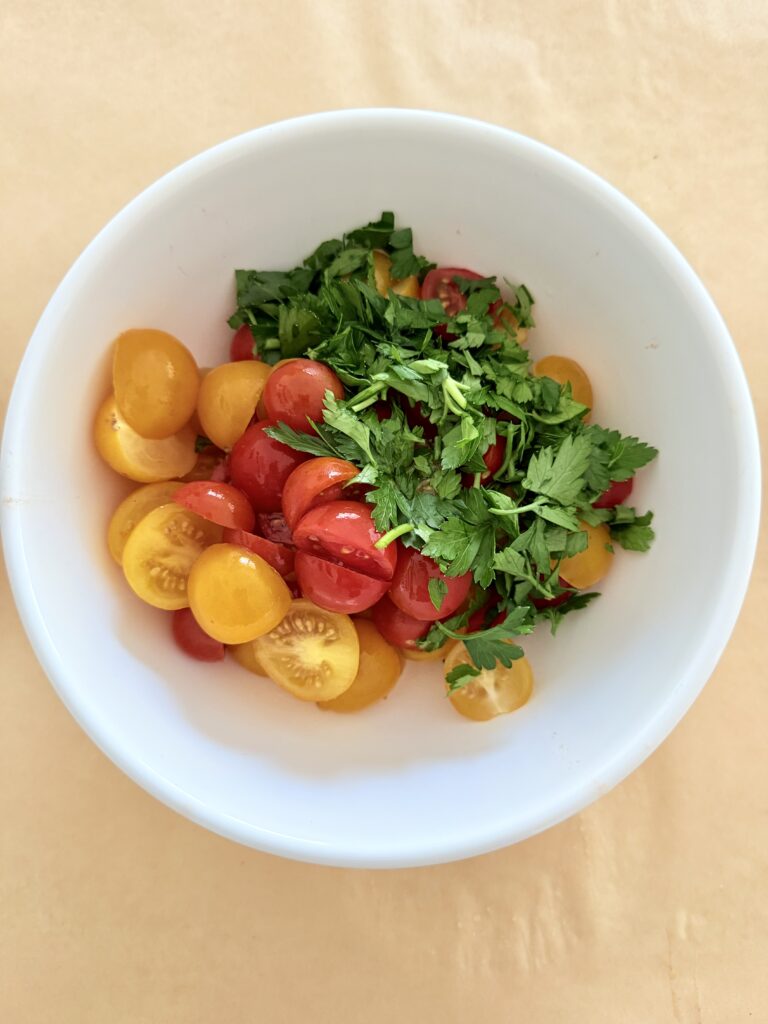 Halved yellow and red cherry tomatoes with chopped parley in a white mixing bowl on parchment paper on the counter.