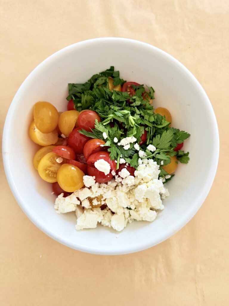 Halved yellow, crumbled feta cheese, red cherry tomatoes with chopped parley in a white mixing bowl on parchment paper on the counter.