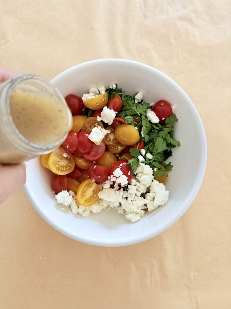 Halved yellow, crumbled feta cheese, red cherry tomatoes with chopped parley in a white mixing bowl on parchment paper on the counter. with greek salad dressing in a small glass container is being poured over the salad ingredients.