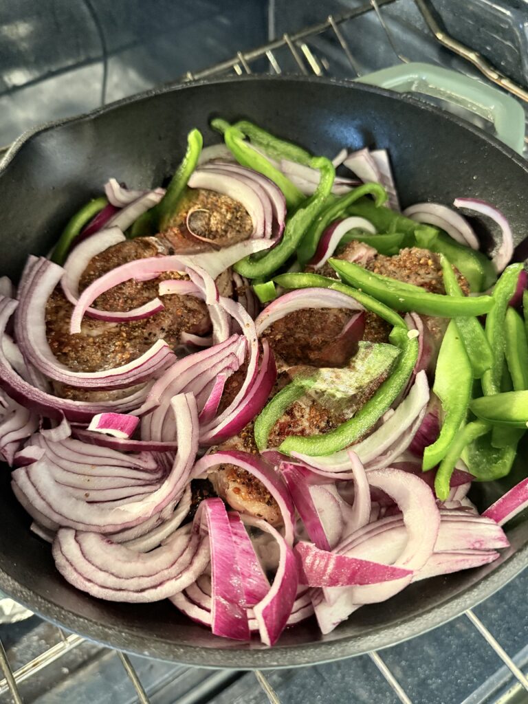 Seared steak in a cast-iron skillet on the stove top with raw sliced red onion and sliced green pepper on top.