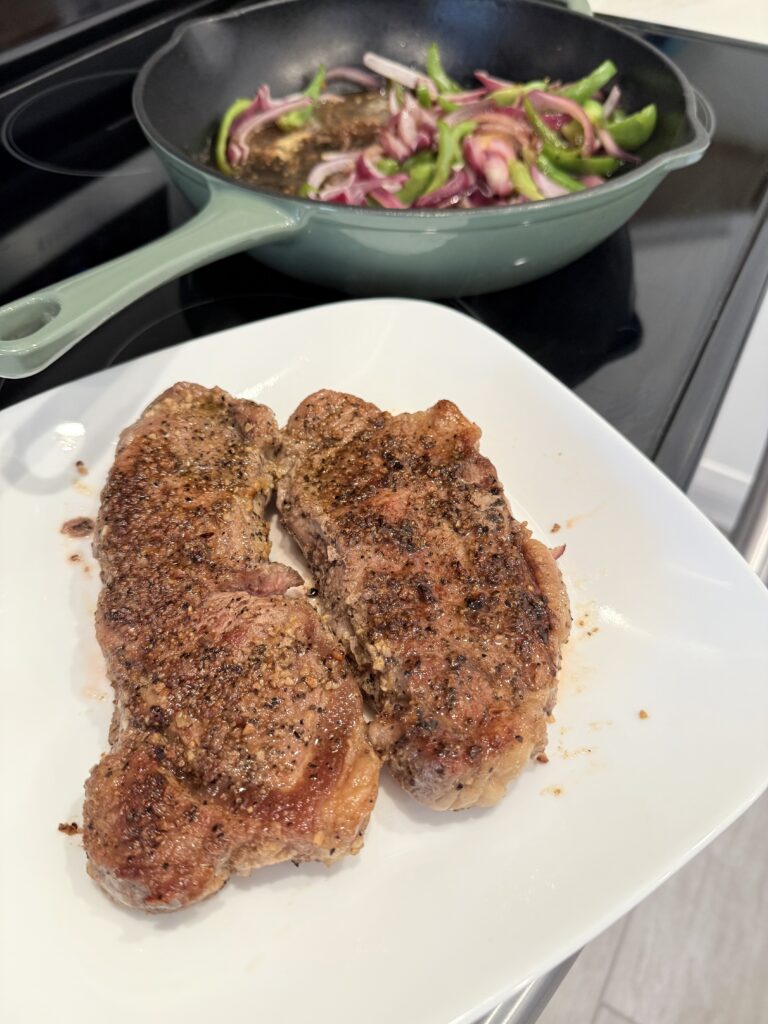 cooked steak on a white plate beside the vegetables in the cast-iron skillet ontop of the stove.