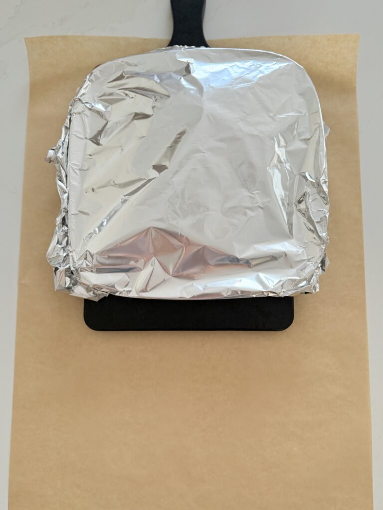 steak resting covered with foil on a plate on a black cutting board on parchment paper on the counter,