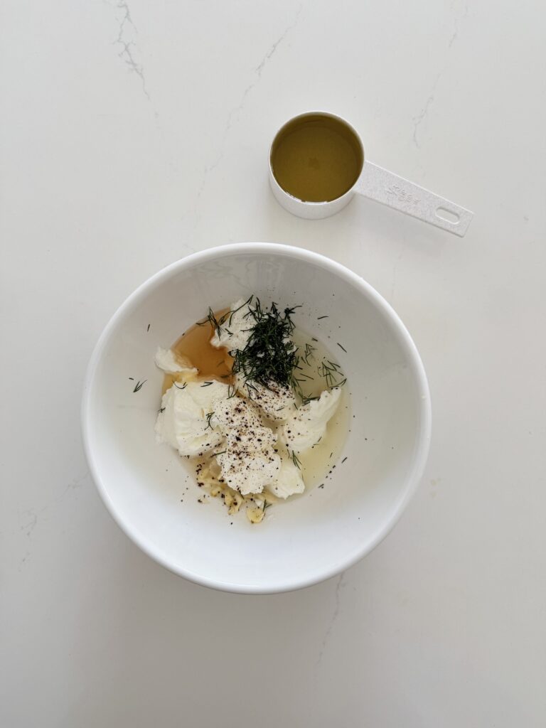 All the ingredients together to make creamy Greek yogurt dressing in a white mixing bowl, except the olive oil in a measuring cup, are all on the counter.