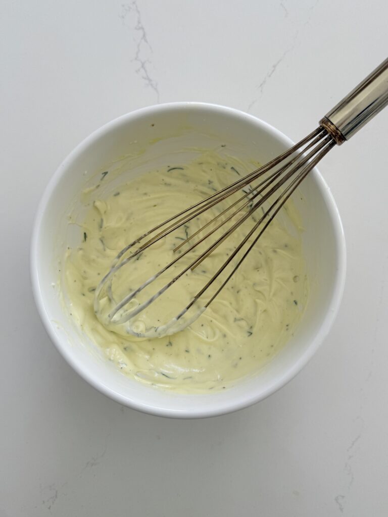 A whisk whisk all of the ingredients together to make creamy Greek yogurt dressing in a white mixing bowl on the counter.