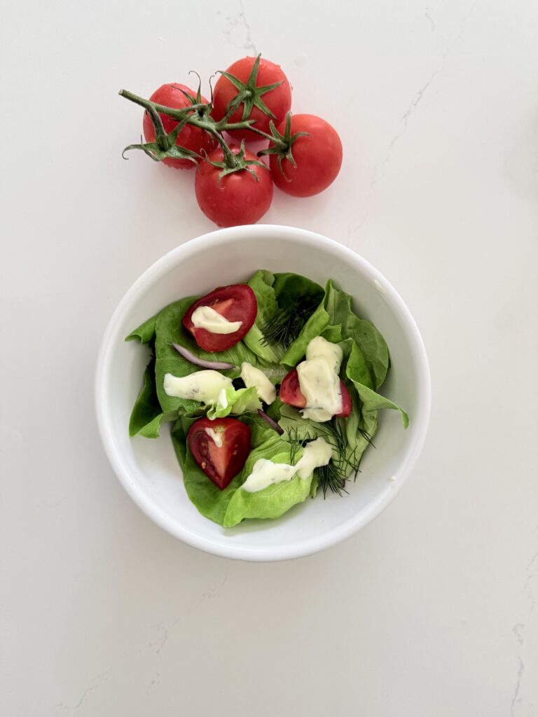 Creamy Greek yogurt dressing on top of butter, lettuce, tomatoes, and a white bowl with cherry tomatoes beside it above the bowl. All on the counter.
