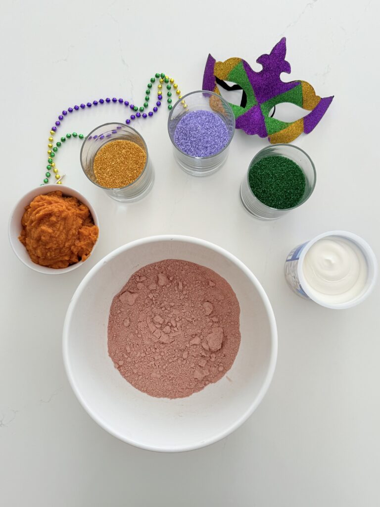 Devil's food cake in a large and white mixing bowl on the counter, canned pumpkin and a small white bowl, white frosting, green, purple, and gold sprinkles in individual glass cups with Mardi Gras decorations on the counter.