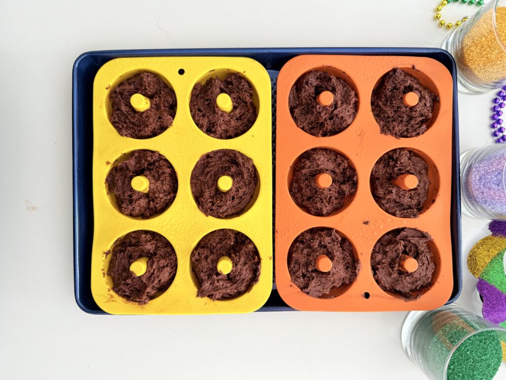 Batter and donut molds on a dark blue baking sheet on the counter, ready to go in the oven. Surrounded by Mardi Gras, colored sprinkles and decorations.