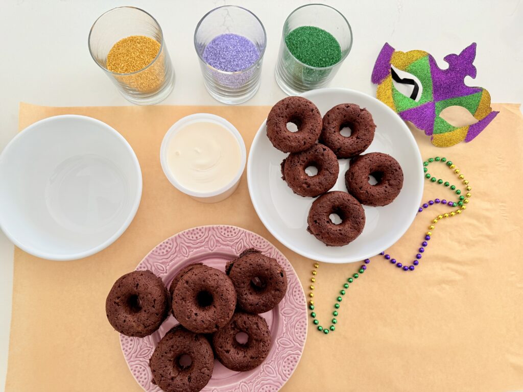 Cool donuts on a white plate and a purple plate on brown parchment paper surrounded by white icing, gold, purple, green sprinkles, and Mardi Gras decorations, all on the counter.