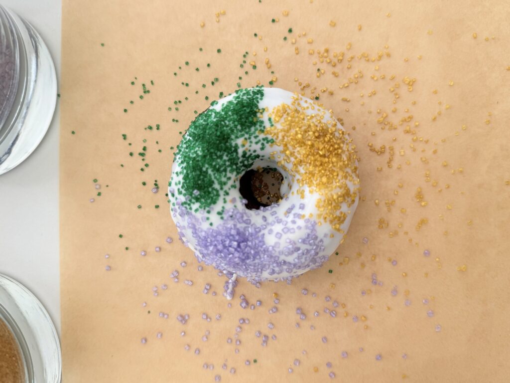 Garnished donut with green, gold, and purple sprinkles on the frosted donut immediately after dipping it into white icing. It sits on brown parchment paper on the counter.