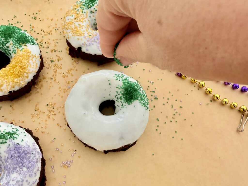 Garnishing the donut with green sprinkles onto the frosted donut immediately after dipping it into white icing. It sits on brown parchment paper on the counter.